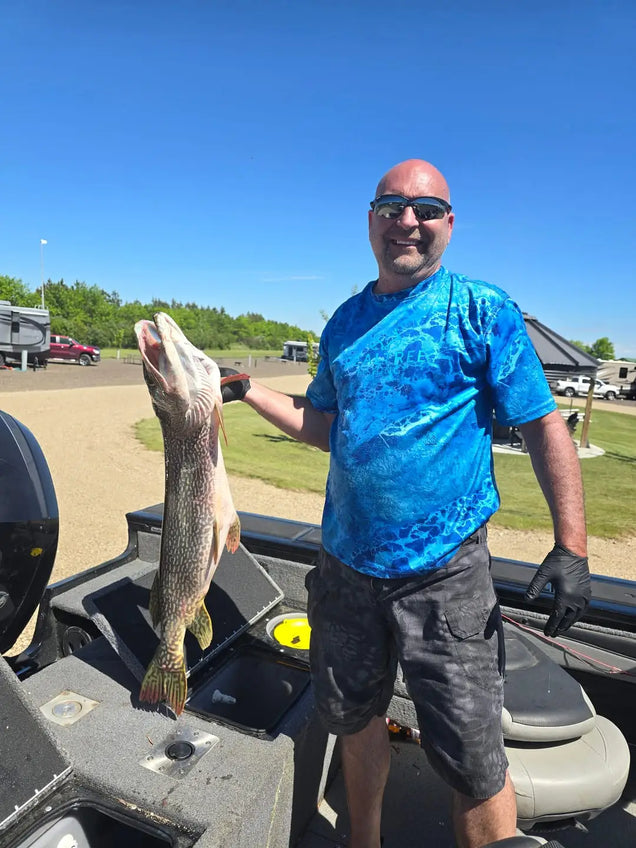 Man holding a large fish on a boat with a clear blue sky and grassy area in the background.