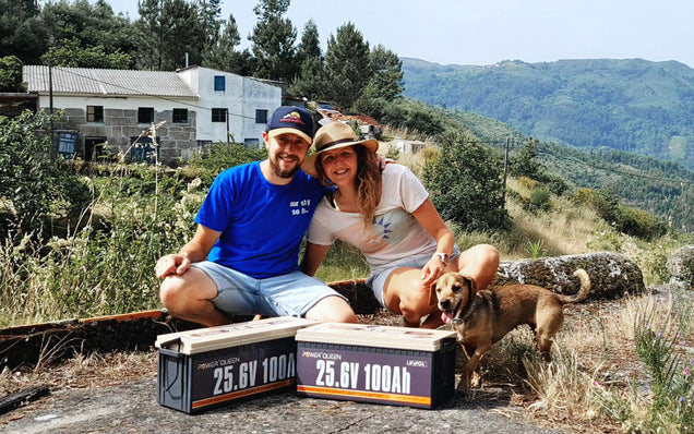 Two people and a dog sitting on a log with two large Power Queen lithium batteries in a scenic outdoor setting.